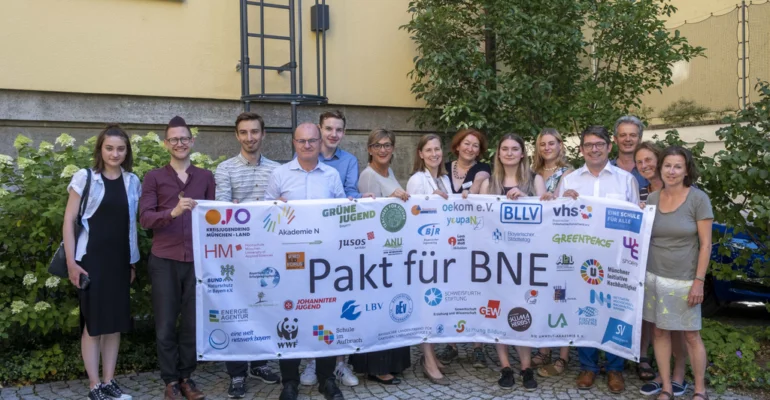 Viele Menschen halten ein Banner mit bunten Logos der Unterzeichner*innen des Pakts für Bildung für nachhaltige Entwicklung in Bayern darauf hoch.